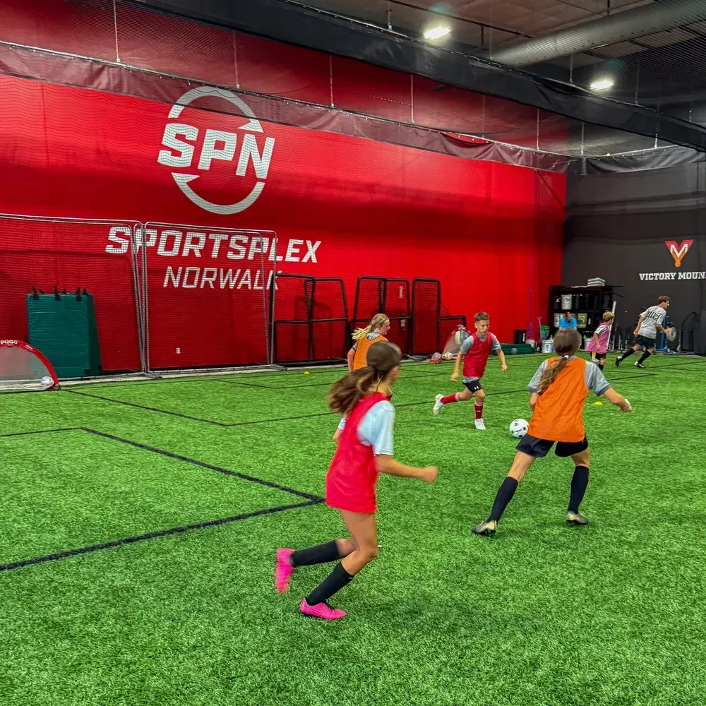 Children playing soccer indoors on green turf at SPN Sportsplex Norwalk, with red wall and nets in the background