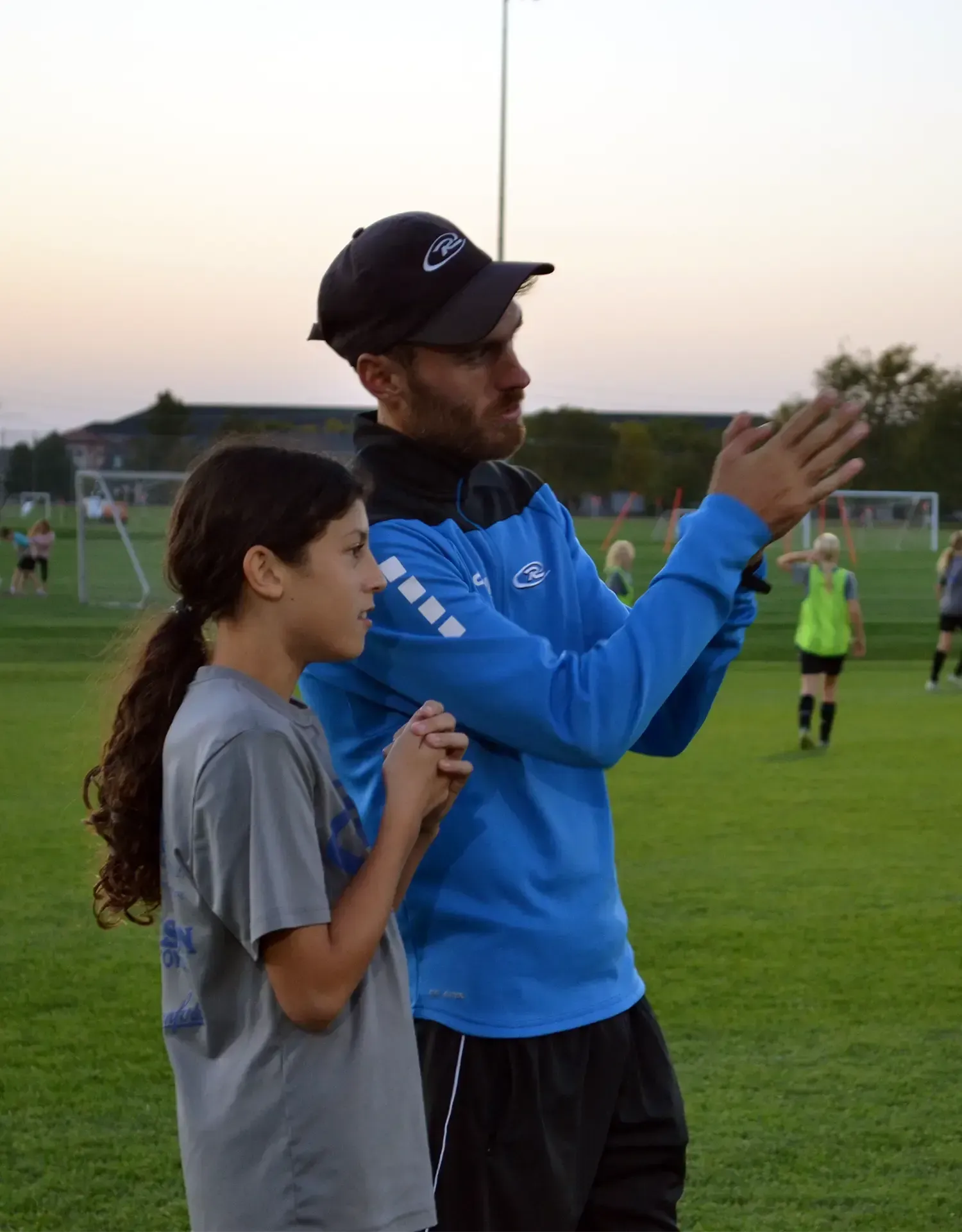 Coach in blue gesturing beside a child on a soccer field at dusk