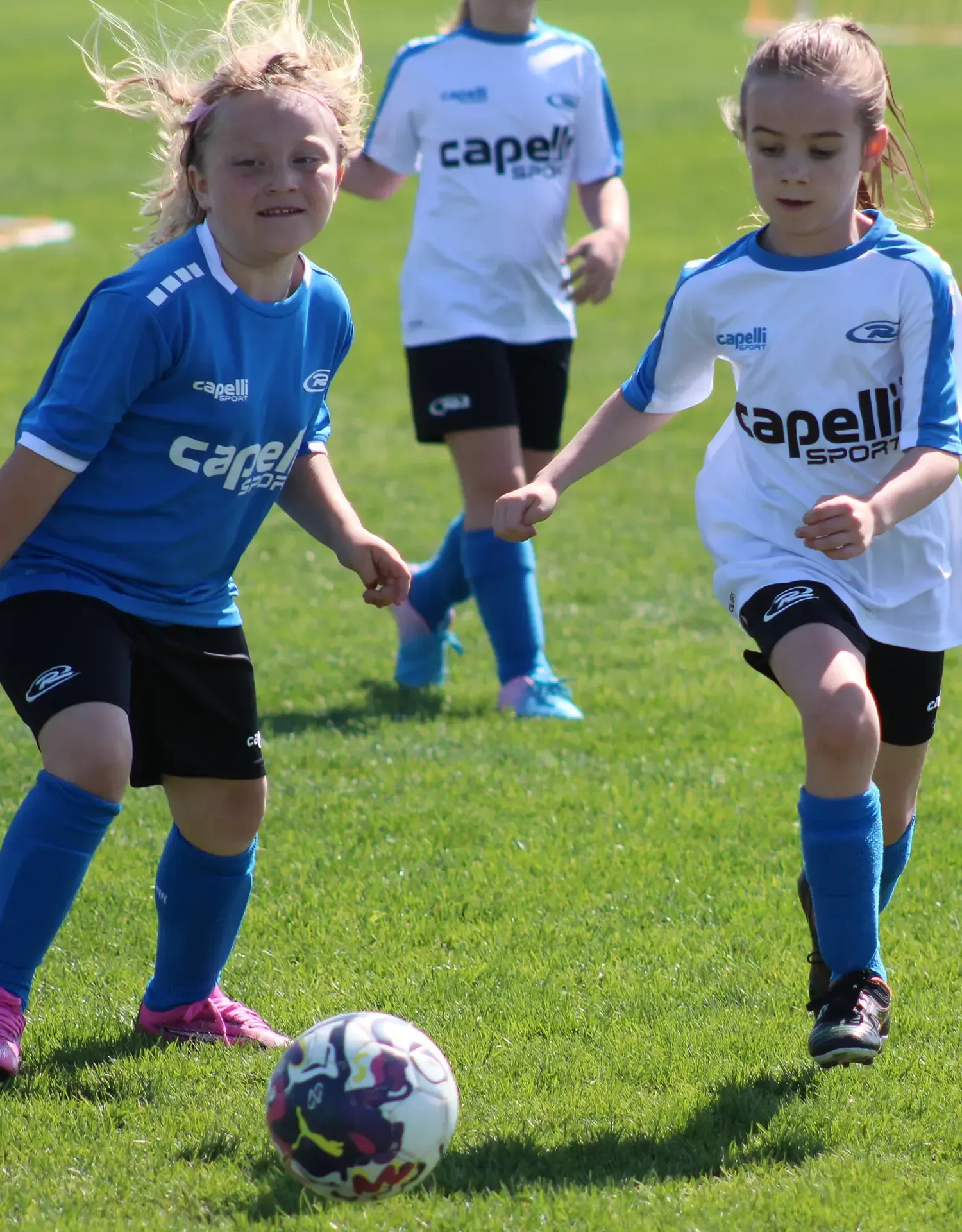Two children in soccer uniforms chasing a ball on a grassy field during a game