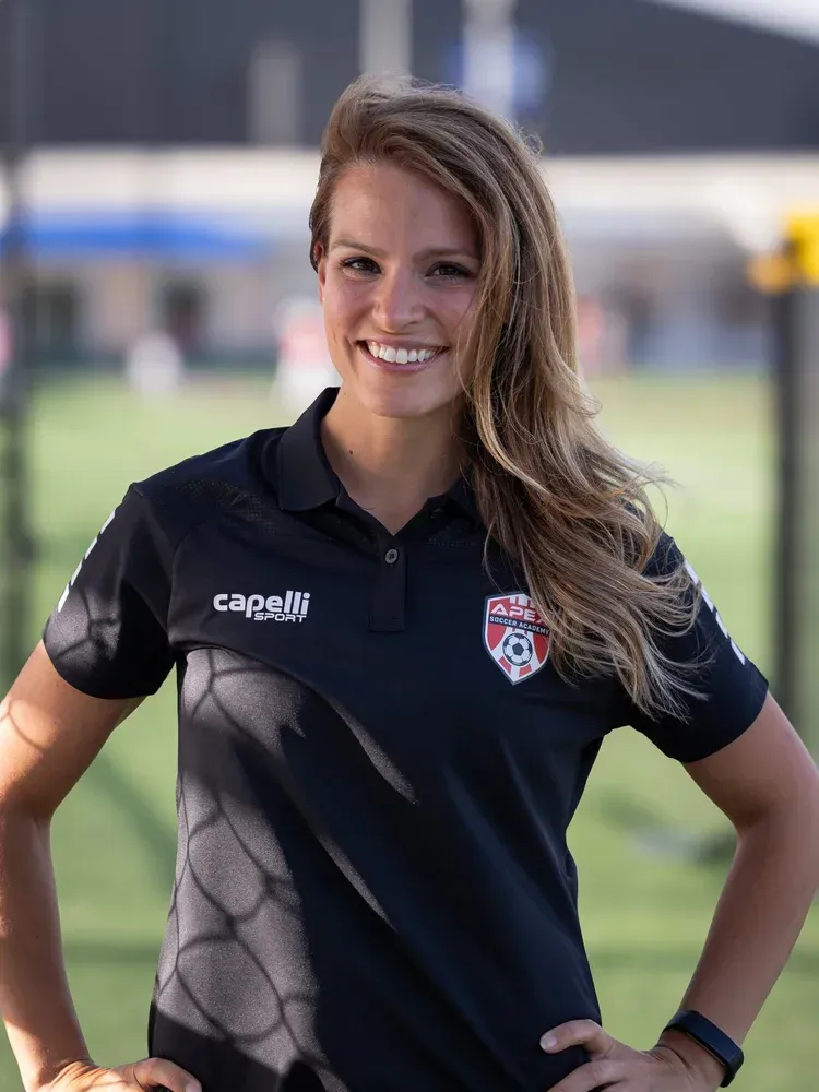 Woman in a black polo shirt smiling outdoors on a soccer field, wearing a team crest.