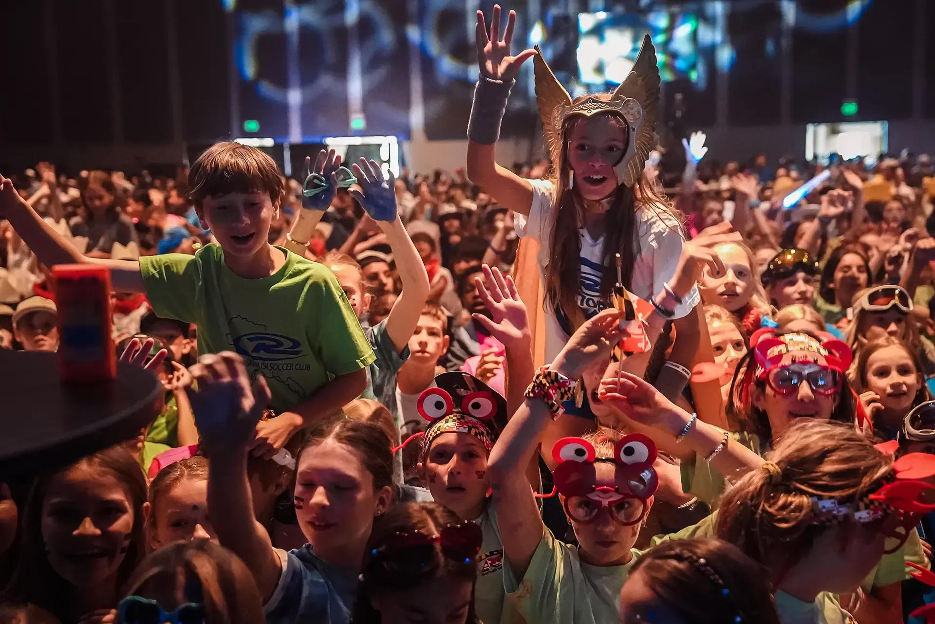 Crowd of children cheering at a colorful indoor event, with lights and raised hands.