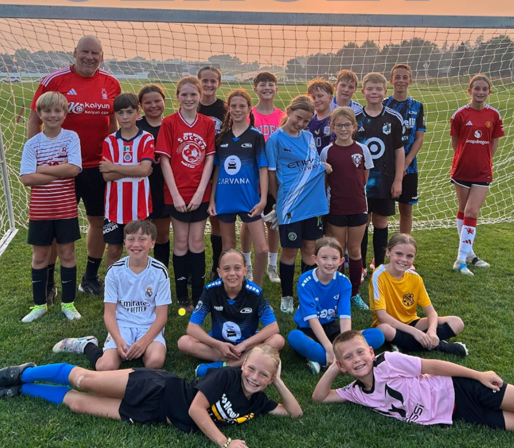 Youth soccer team posing on a field at sunset, wearing red, blue, and black jerseys.