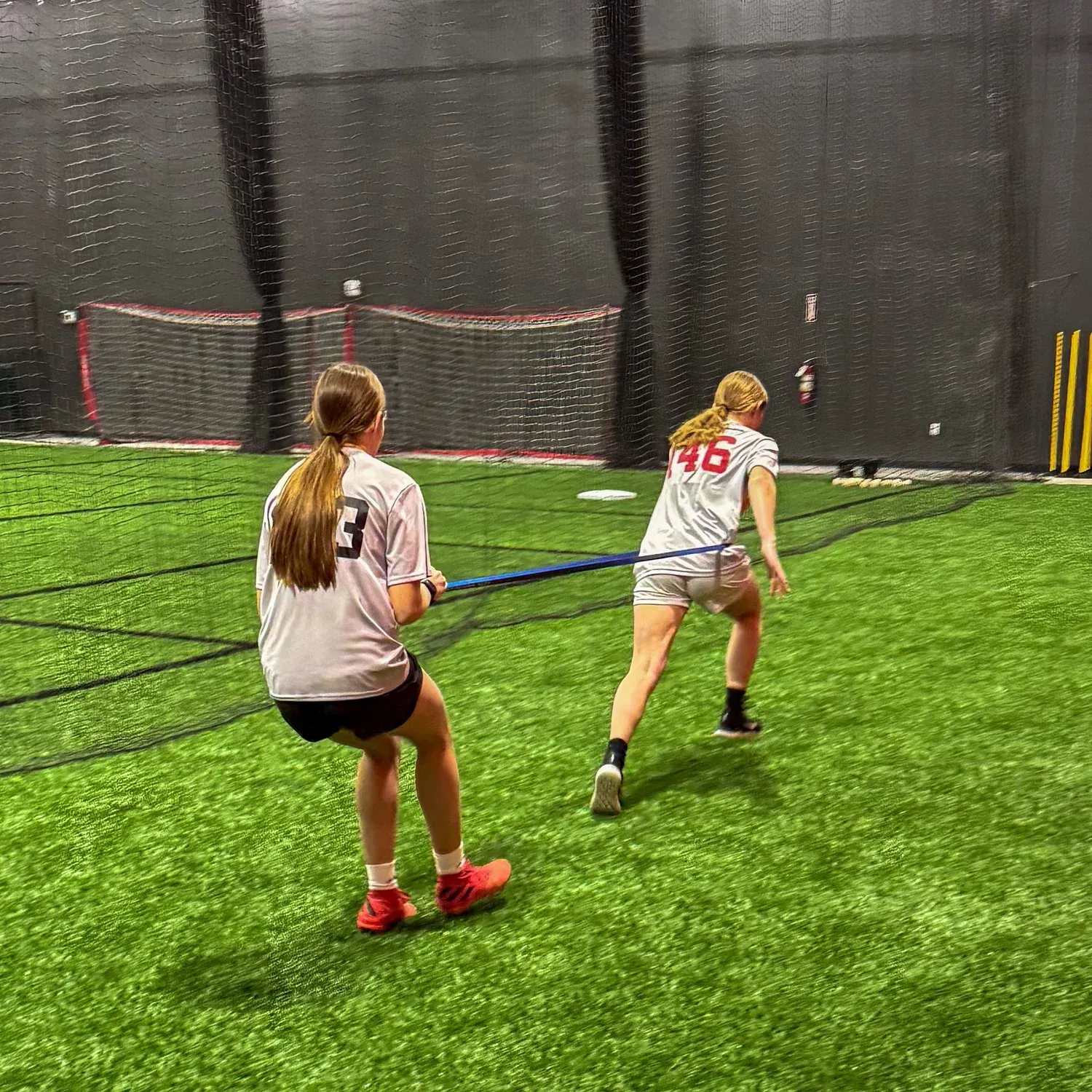 Two athletes in white jerseys on indoor turf, poised to move near a goal net.