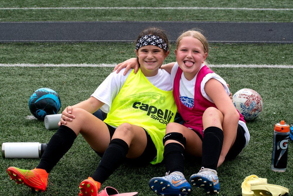 Two soccer players in neon jerseys sit on turf, smiling with sports balls and water bottles nearby.