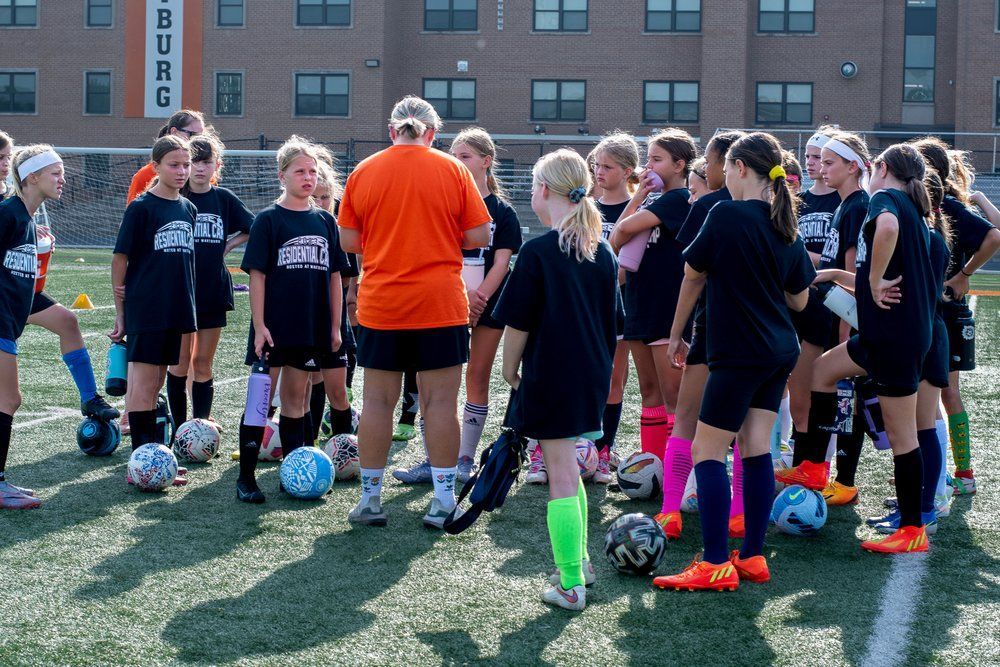 Youth soccer team huddled with coach on a field, holding blue soccer balls in front of a building