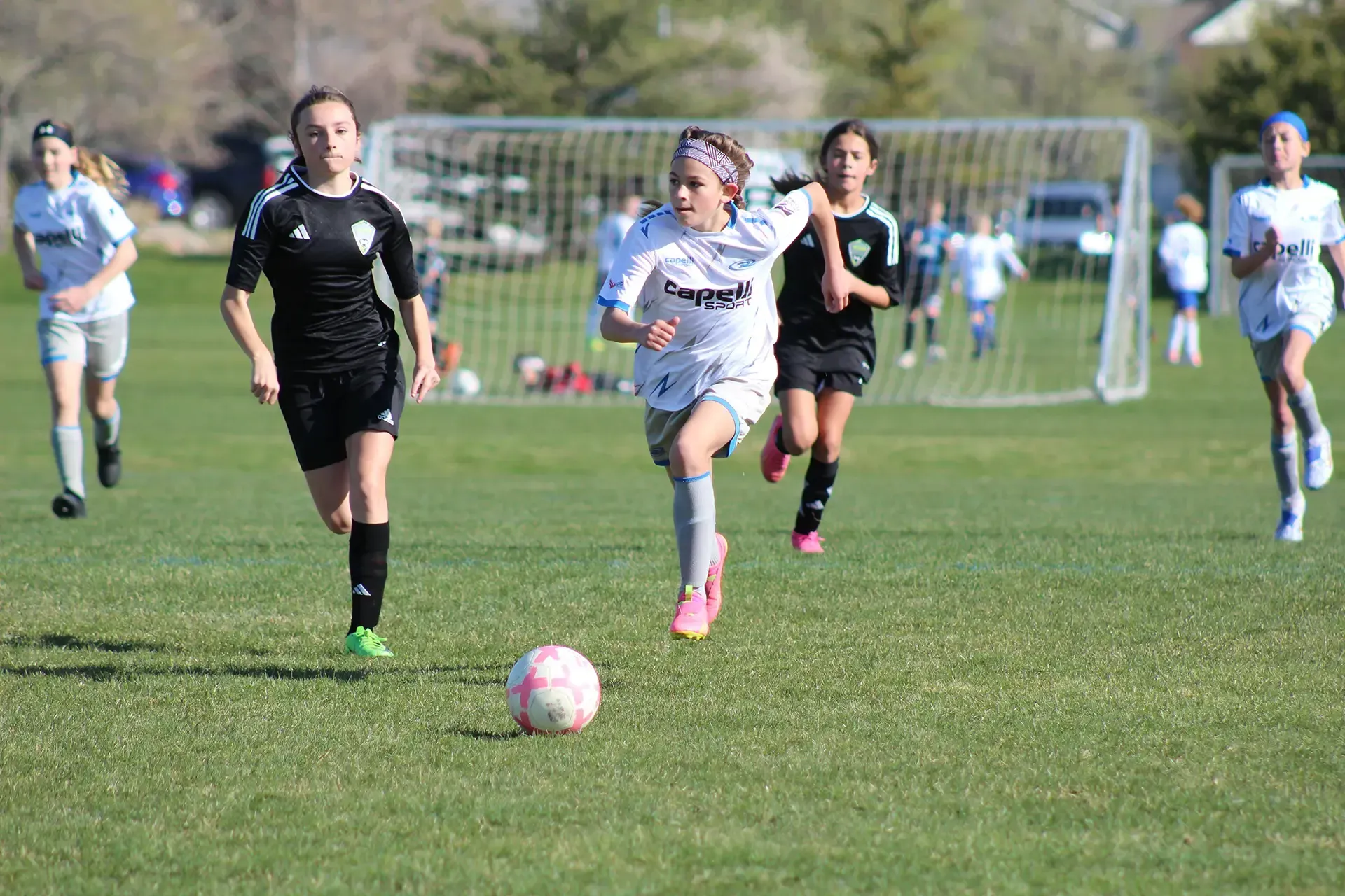 Soccer players sprint toward the ball on a sunny field, with a goal visible in the background.