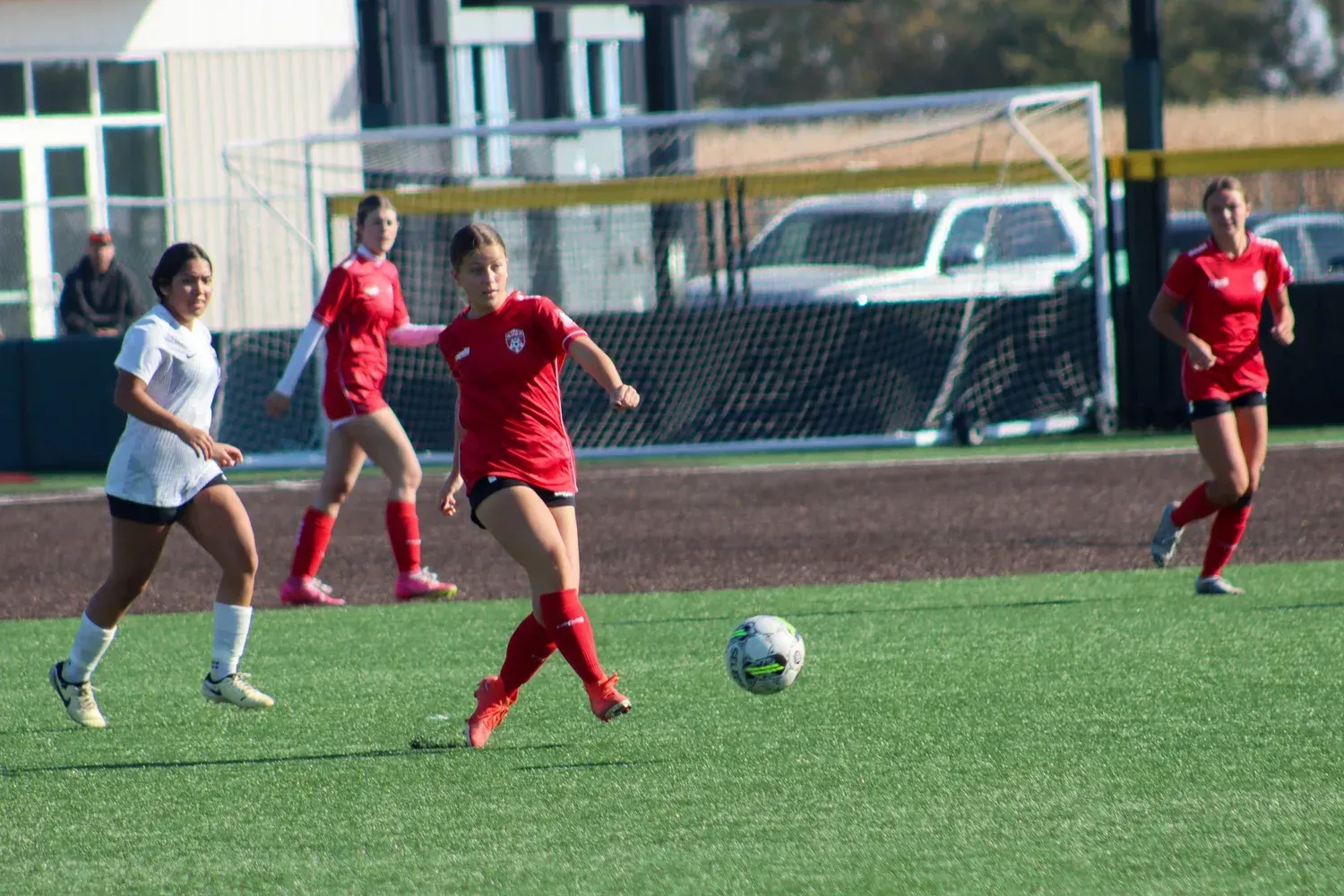 Soccer players in red and white jerseys chase the ball on a grassy field