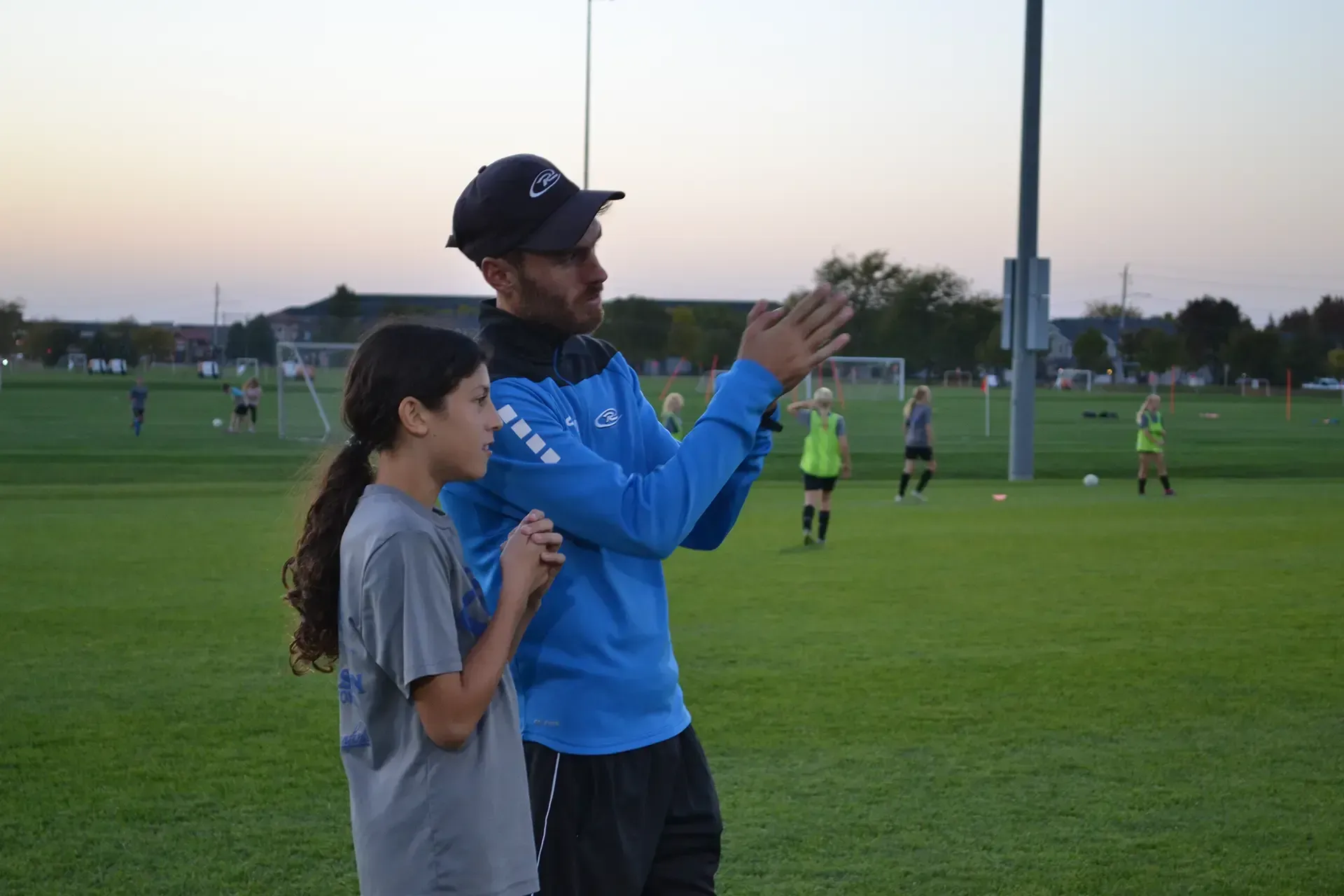 Two people in athletic wear stand on a grassy sports field at dusk, watching a game and holding a phone.