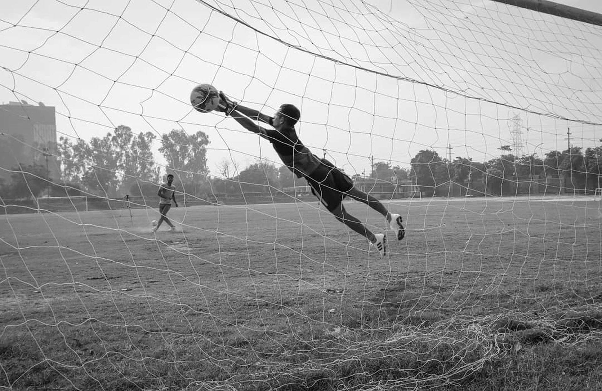 Goalkeeper diving to catch a soccer ball in front of the net on a grassy field.