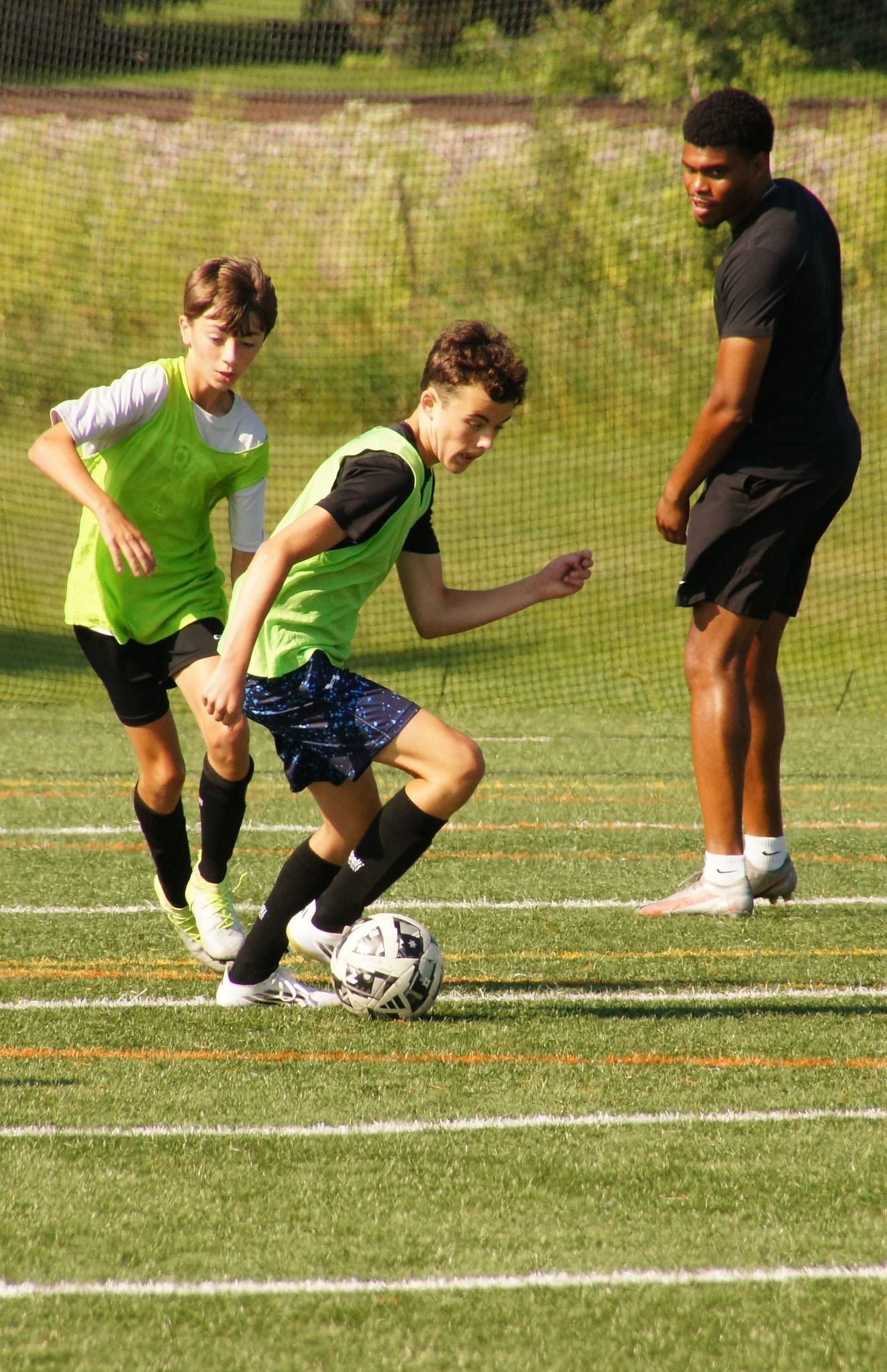 Two soccer players contest the ball while a referee stands nearby on a field.