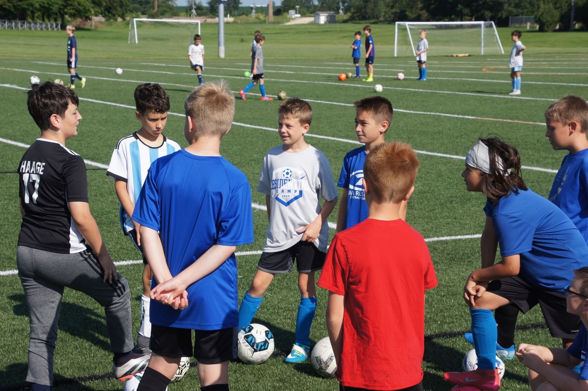 Children in soccer jerseys gather around a ball on a grassy field during practice.