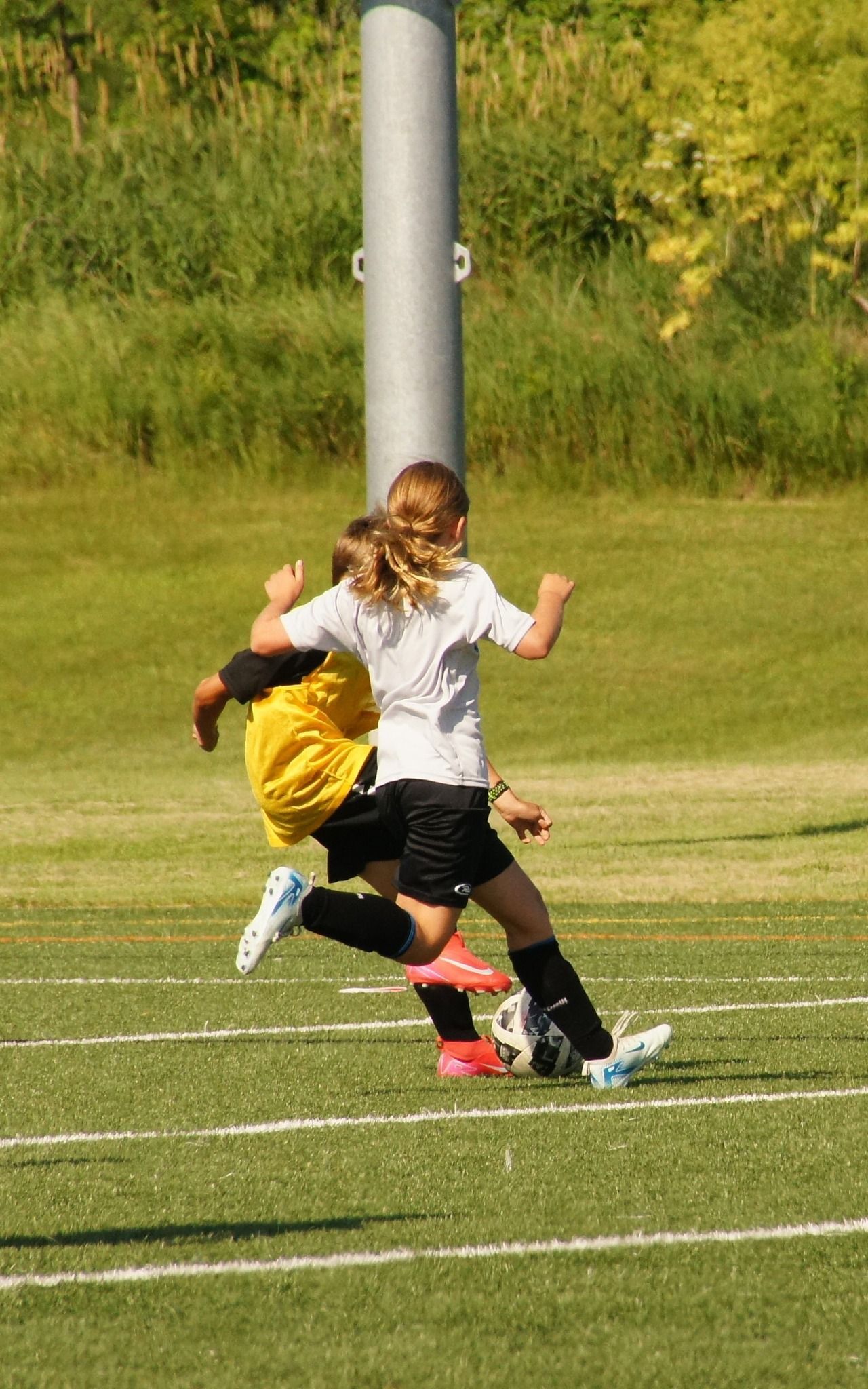 Two children collide on a grassy soccer field, one in white and black, the other in yellow, with a goal post behind them