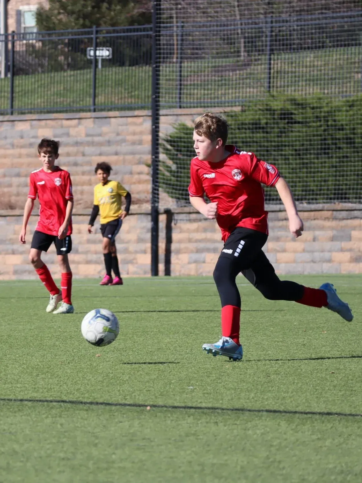 Soccer player in red dribbles a ball on a field while teammates and a referee stand behind him