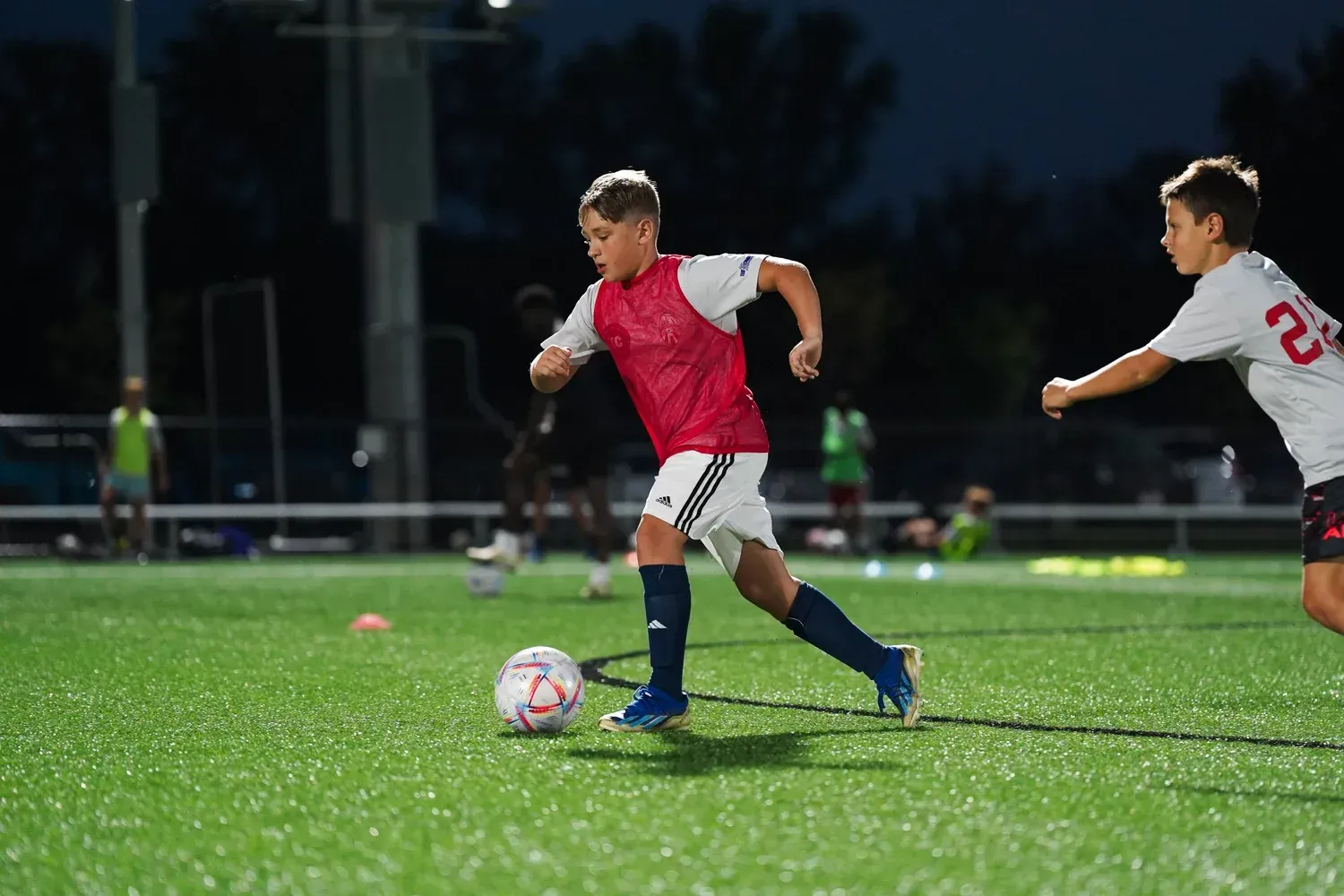 Two boys in red soccer jerseys running on a lit field, chasing a ball at night