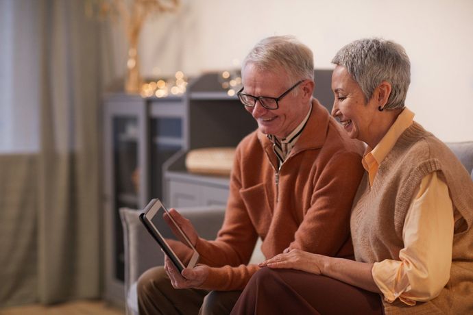 An elderly couple is sitting on a couch looking at a tablet.