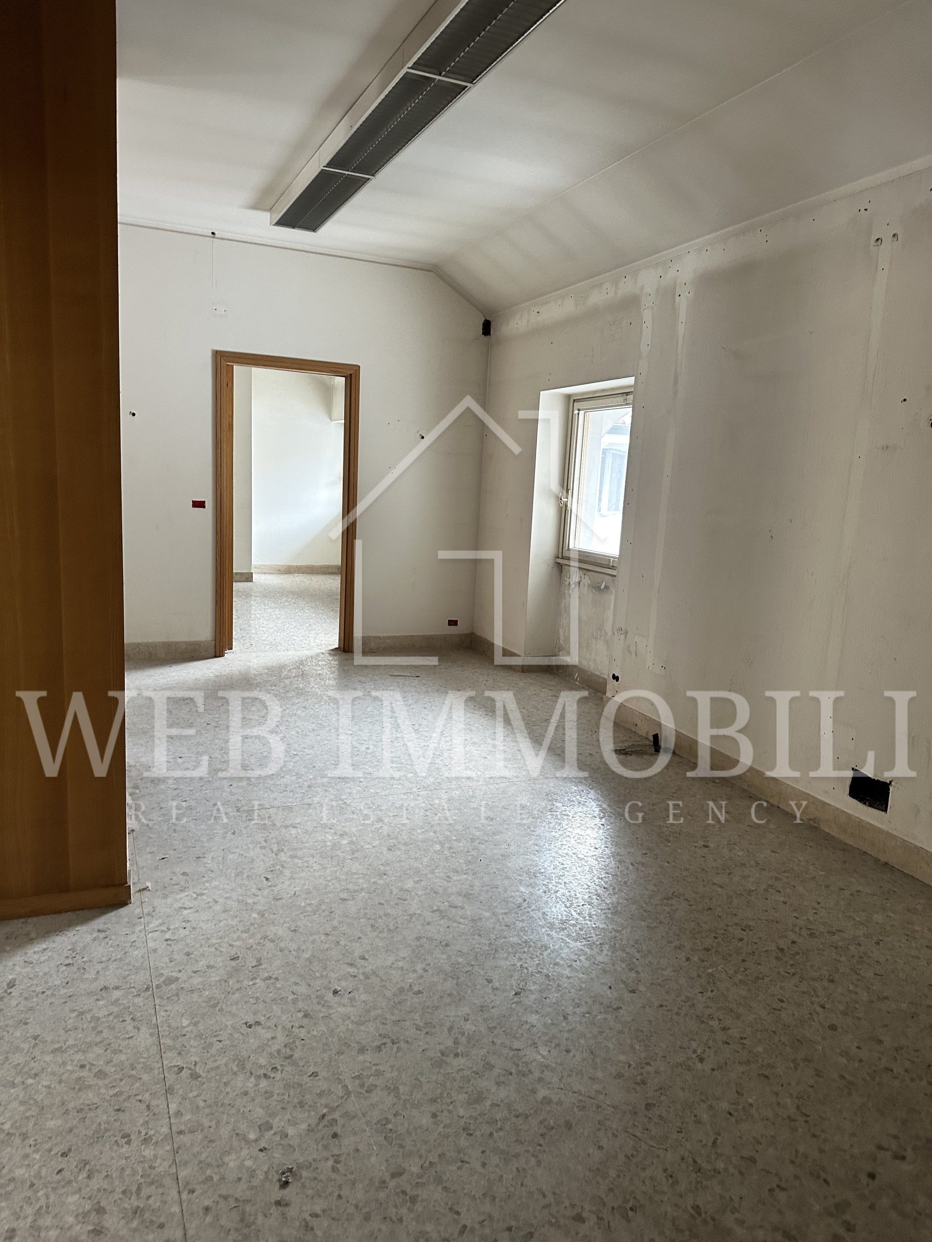Stanza vuota con pavimento in terrazzo, porta che conduce a un'altra stanza, finestra con tenda e pareti spoglie.