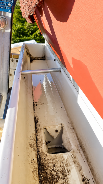 White gutter with debris and a damaged drain pipe beside a red building wall.