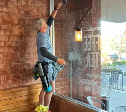 Person on a ladder reaching to clean a hanging lantern on a brick storefront patio.