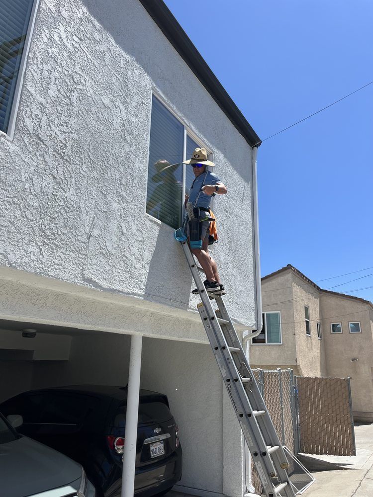 Worker on a ladder painting the white exterior wall of a house above a carport.