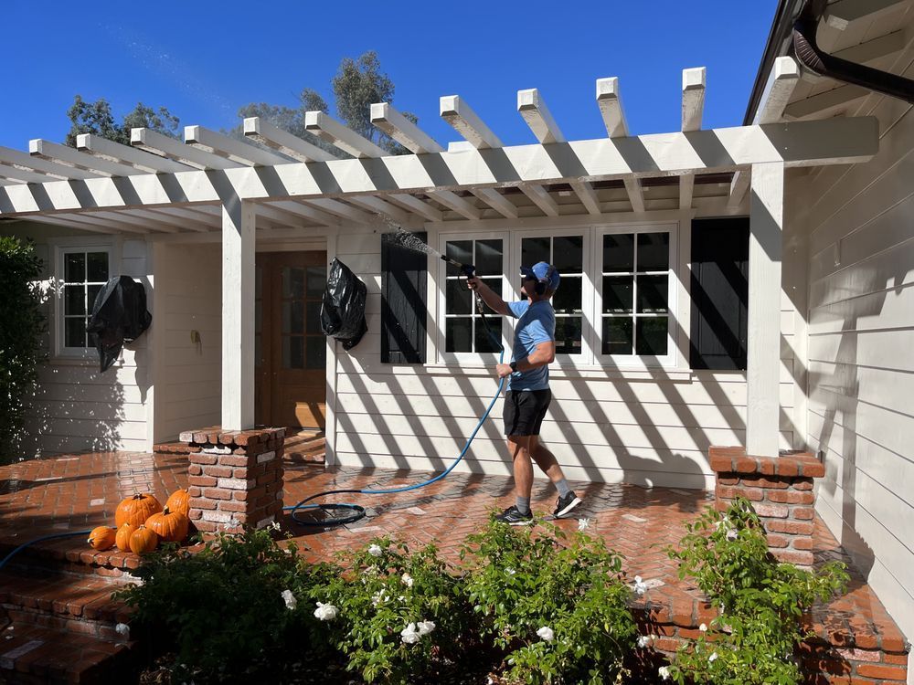 Person painting white pergola beams outside a beige house with brick planter and flowers.