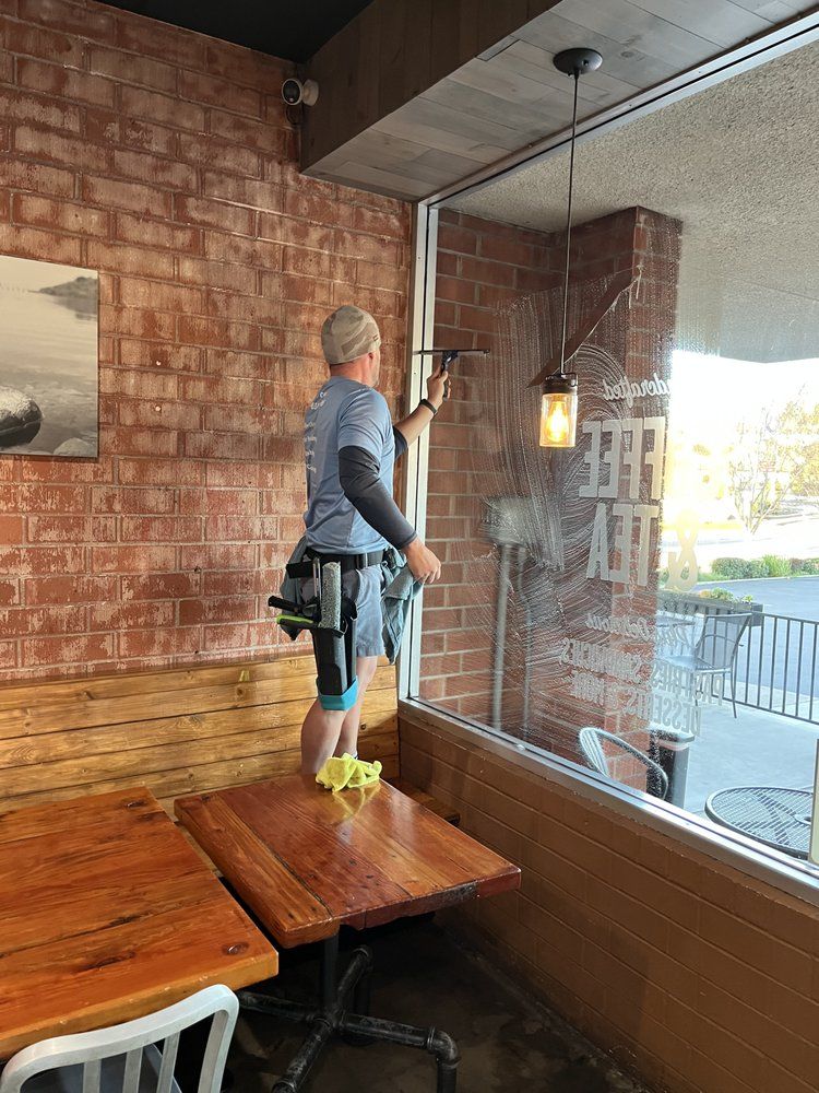 Worker cleaning a large window inside a brick-walled café, standing on a bench and using a squeegee