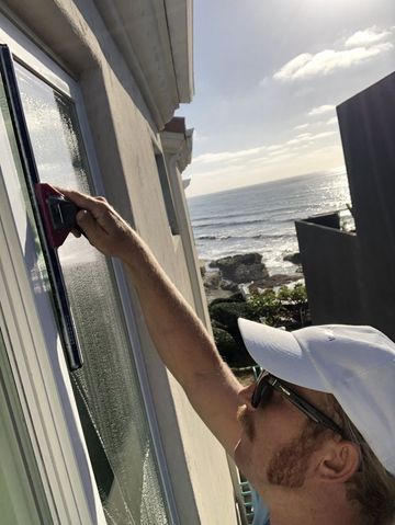 Person cleaning a window with a squeegee, with ocean and rocky shoreline in the background.