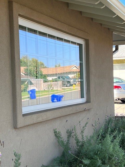 House window with blue tape marks, stucco wall, shrub, and street view reflected in the glass