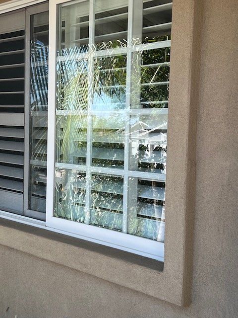 Broken window with shattered glass in a beige stucco wall beside a shuttered window