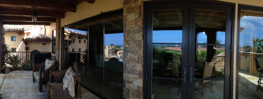 Patio with wicker chairs beside large glass windows, overlooking a sunny garden and blue sky