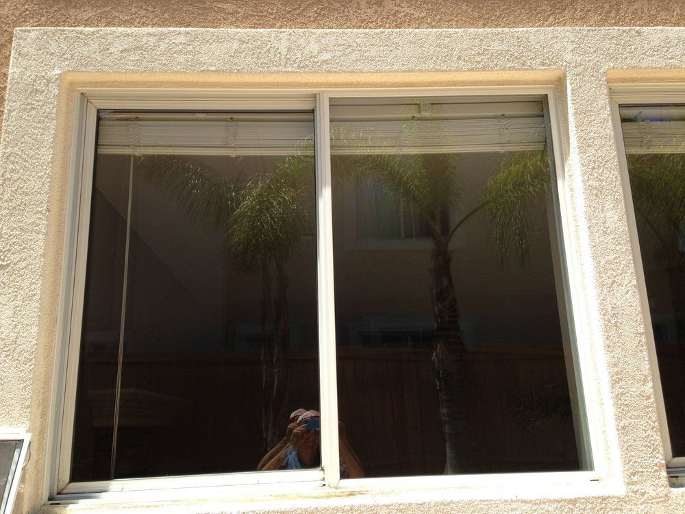 Tan stucco wall with a large window and closed blinds, reflecting palm trees and a person taking the photo