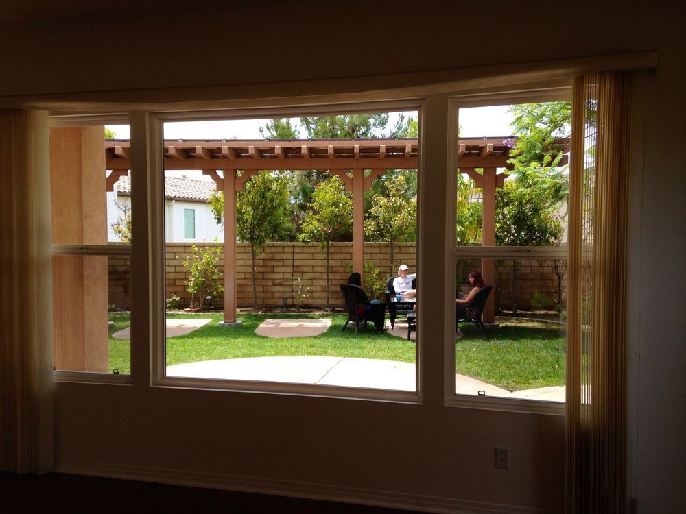 Living room view through a large window to a sunny backyard patio with people seated under a pergola