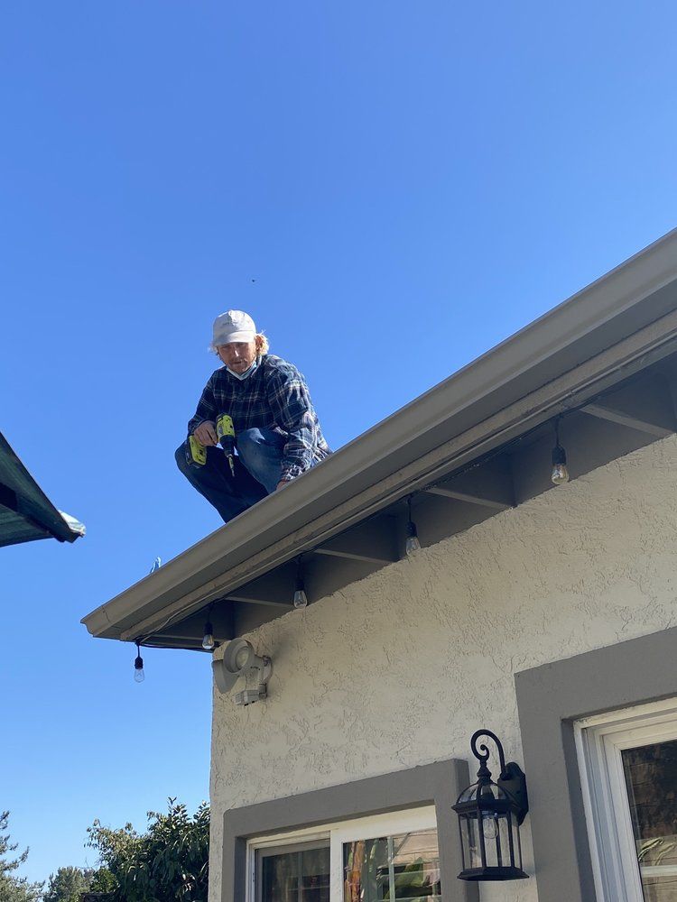 Worker crouched on a roof edge under a blue sky, using a harness and tool belt.