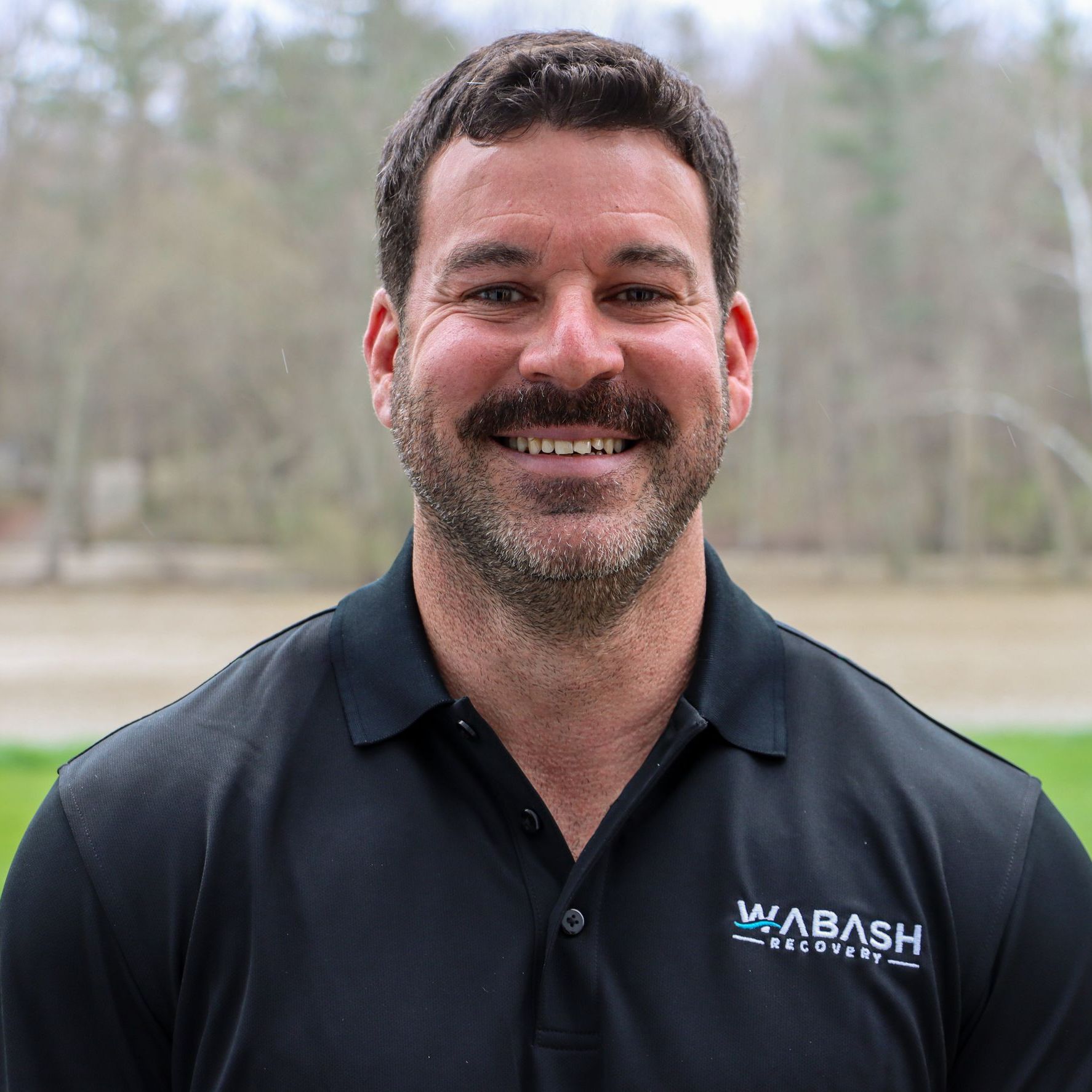 A man with a mustache is wearing a black shirt and smiling.