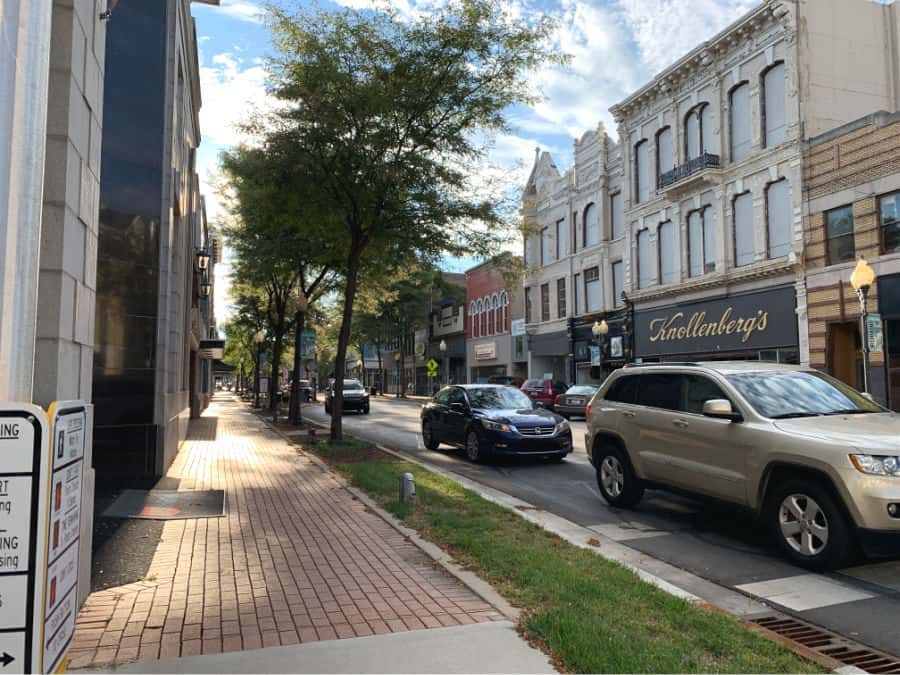 A row of cars are driving down a brick sidewalk in a city.