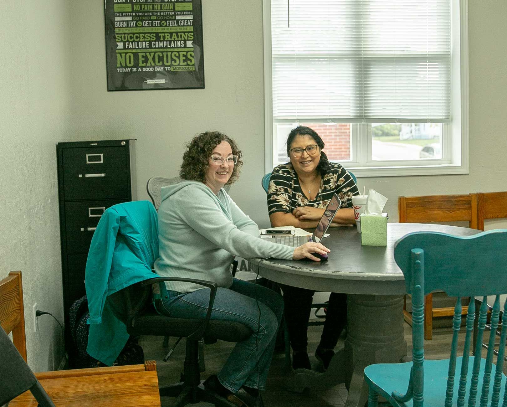 Two women sit at a table in front of a sign that says no excuses