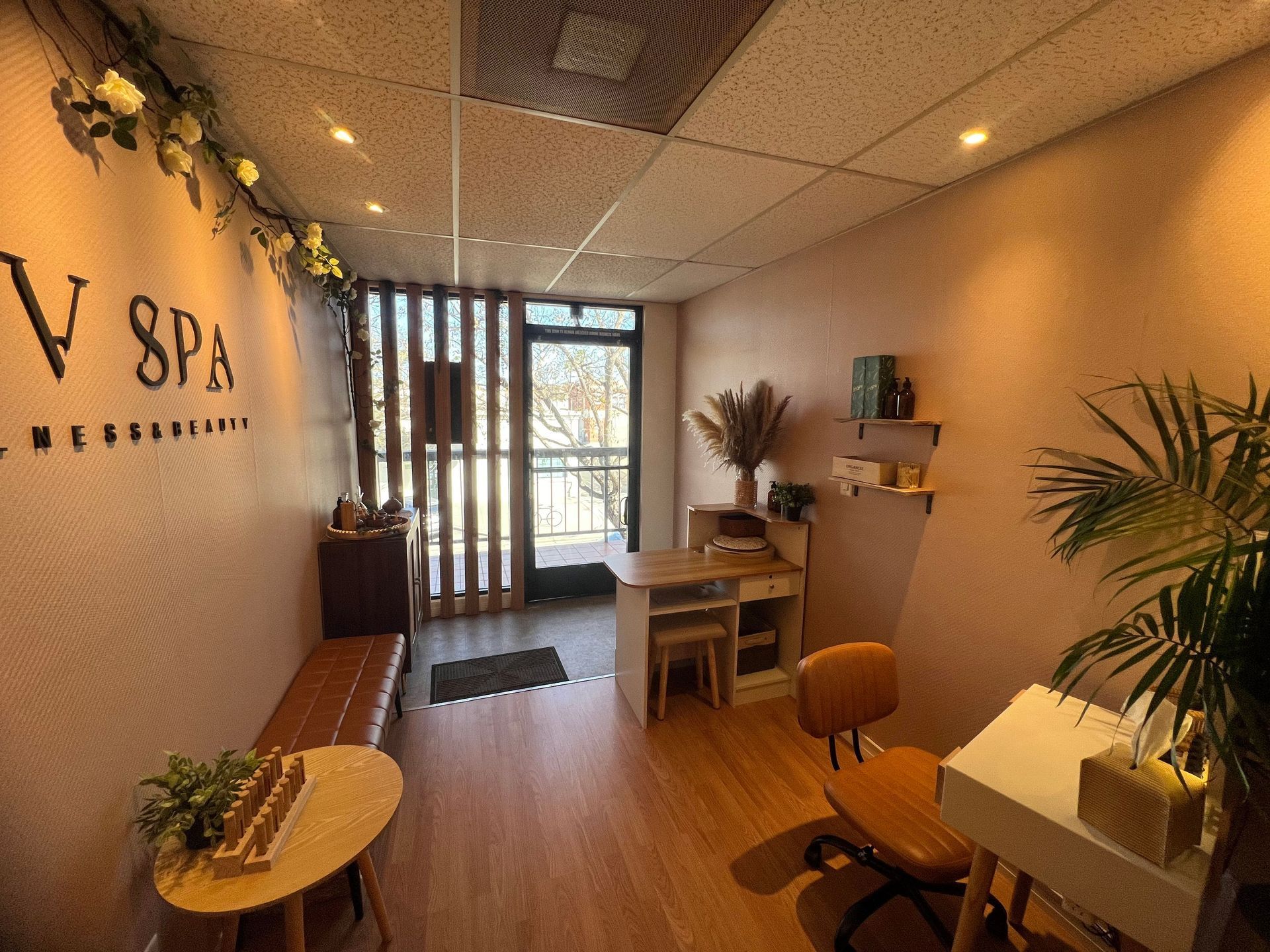Spa interior with pink walls, wooden furniture, and greenery. A desk, bench, and potted plants are visible.