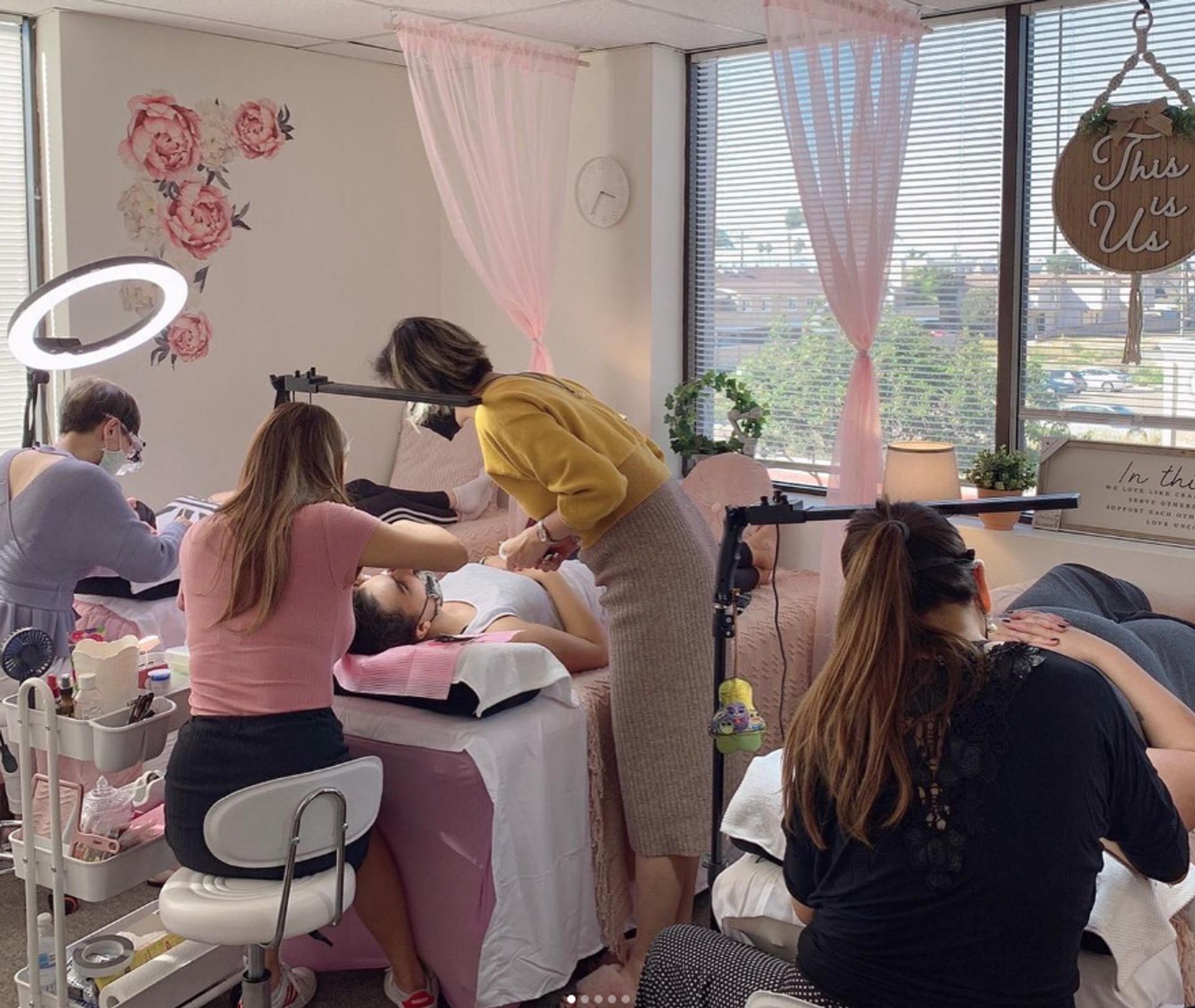 Salon interior with several people applying lash extensions. Pink decor, ring lights, and large windows.