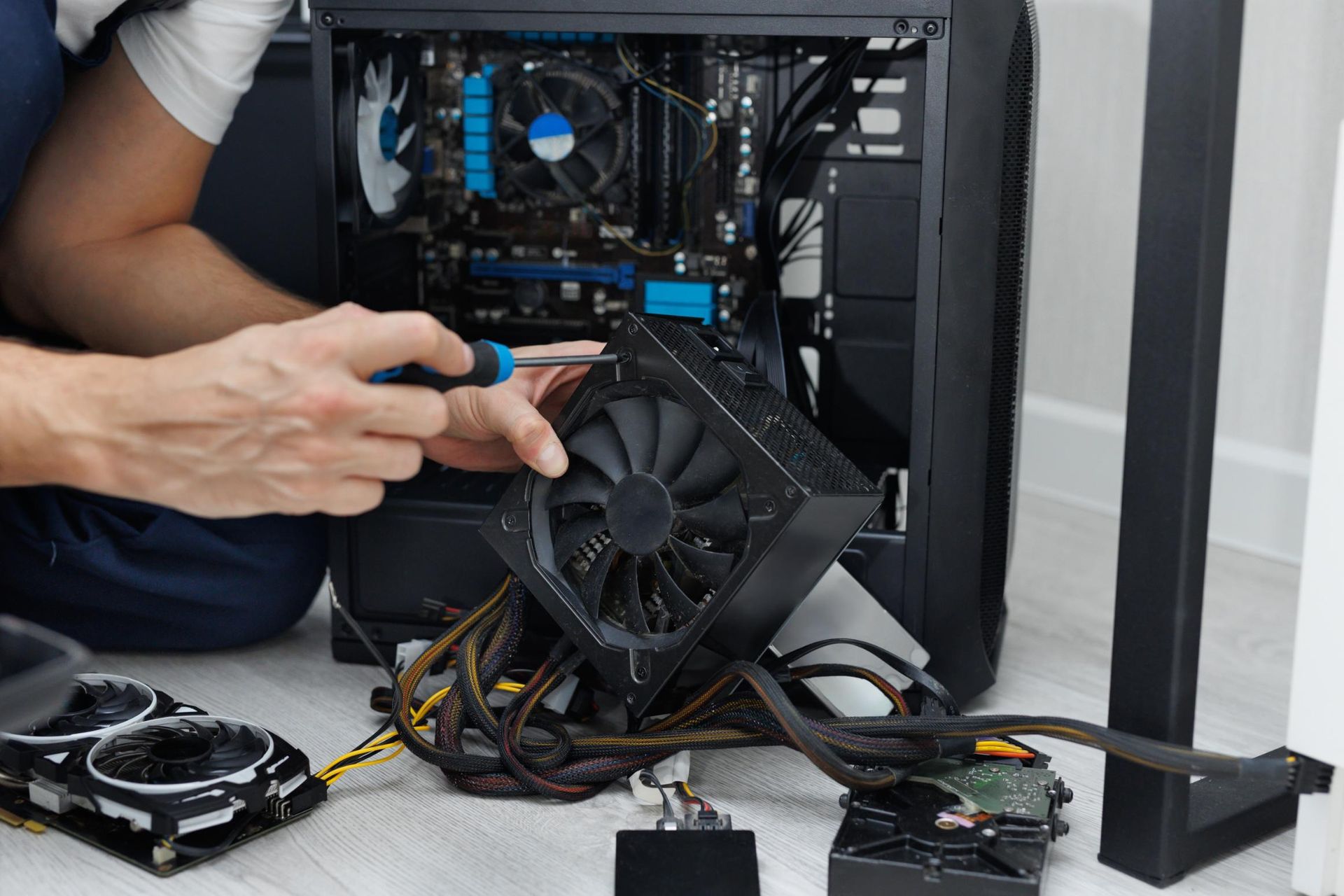 Person disassembles computer, removing a fan with a screwdriver. Various components and cables on the floor.