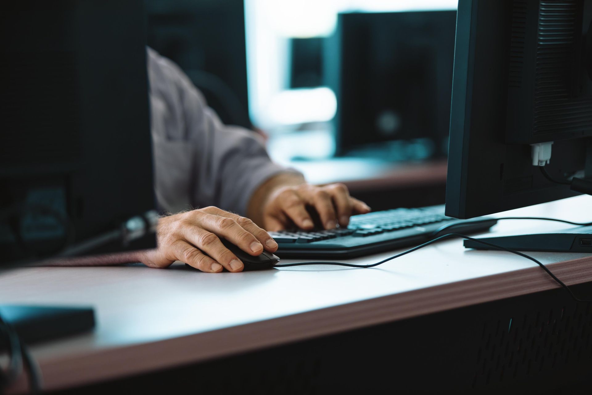 Person's hands on a mouse and keyboard in front of a computer monitor at a desk in an office.