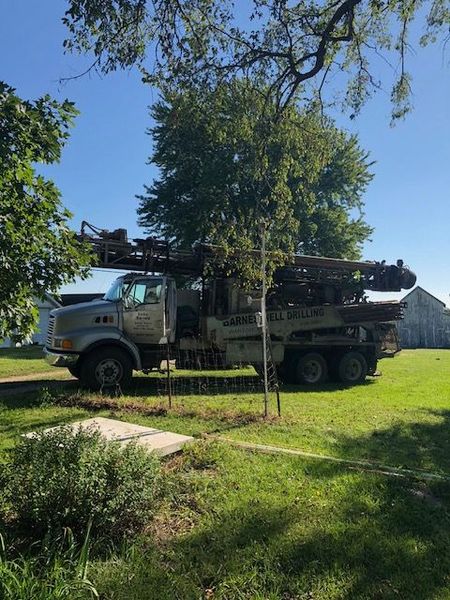 Well drilling truck parked on a grassy lawn with a tree in the background under a blue sky.