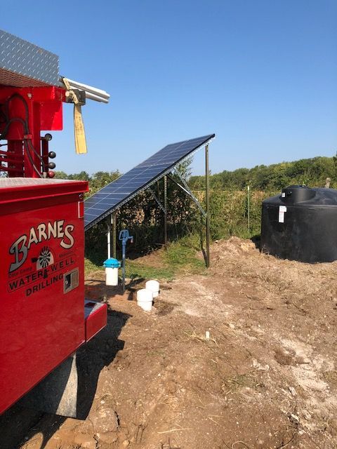 Red truck, solar panels, water well equipment, and black water tank in a field under a blue sky.