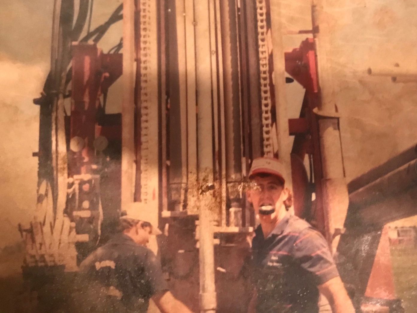 Two men near a drilling rig, likely for a well. One man in a cap holds something in his mouth. Red and beige machine.