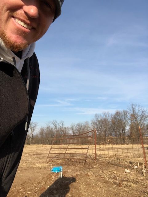 Man smiling, looking at the camera. In a field with a gate and leafless trees under a blue sky.