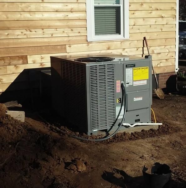 A gray HVAC unit sits on a concrete pad next to the side of a house with light wood siding and freshly dug dirt.
