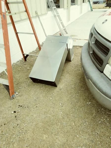 A large, silver rectangular metal duct section sits on a gravel lot next to a ladder and the front bumper of a vehicle.