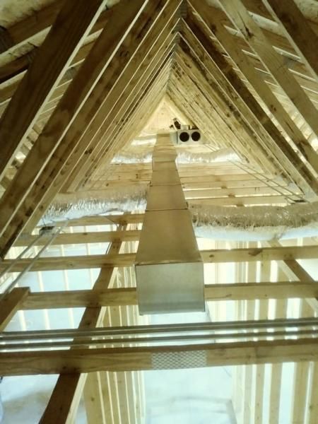 A metal rectangular HVAC duct runs through the center of an unfinished attic with wooden framing and roof trusses.