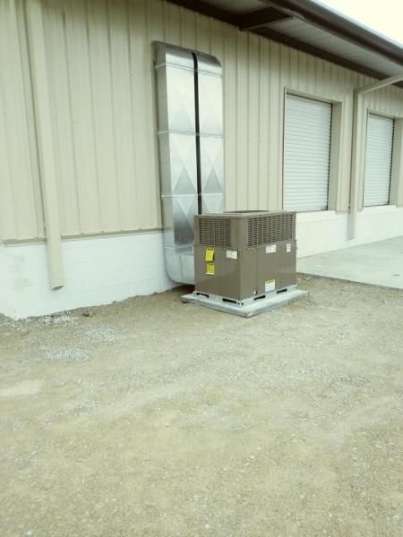 An exterior HVAC unit sits on a concrete pad against a metal-sided building with a vertical metal duct attached to the wall.
