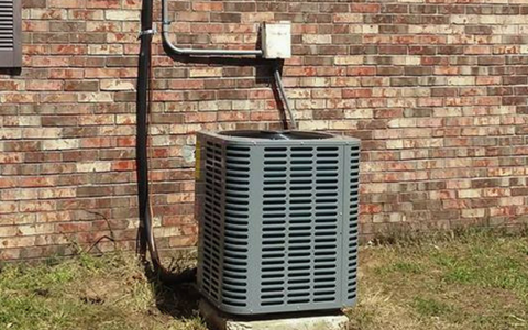 A gray HVAC condenser unit sits on a concrete pad against a brown brick house wall with electrical conduit and wiring.