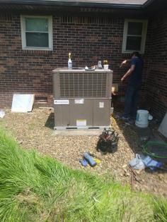 A technician works on an outdoor HVAC unit located on a gravel patch against a brick house wall.
