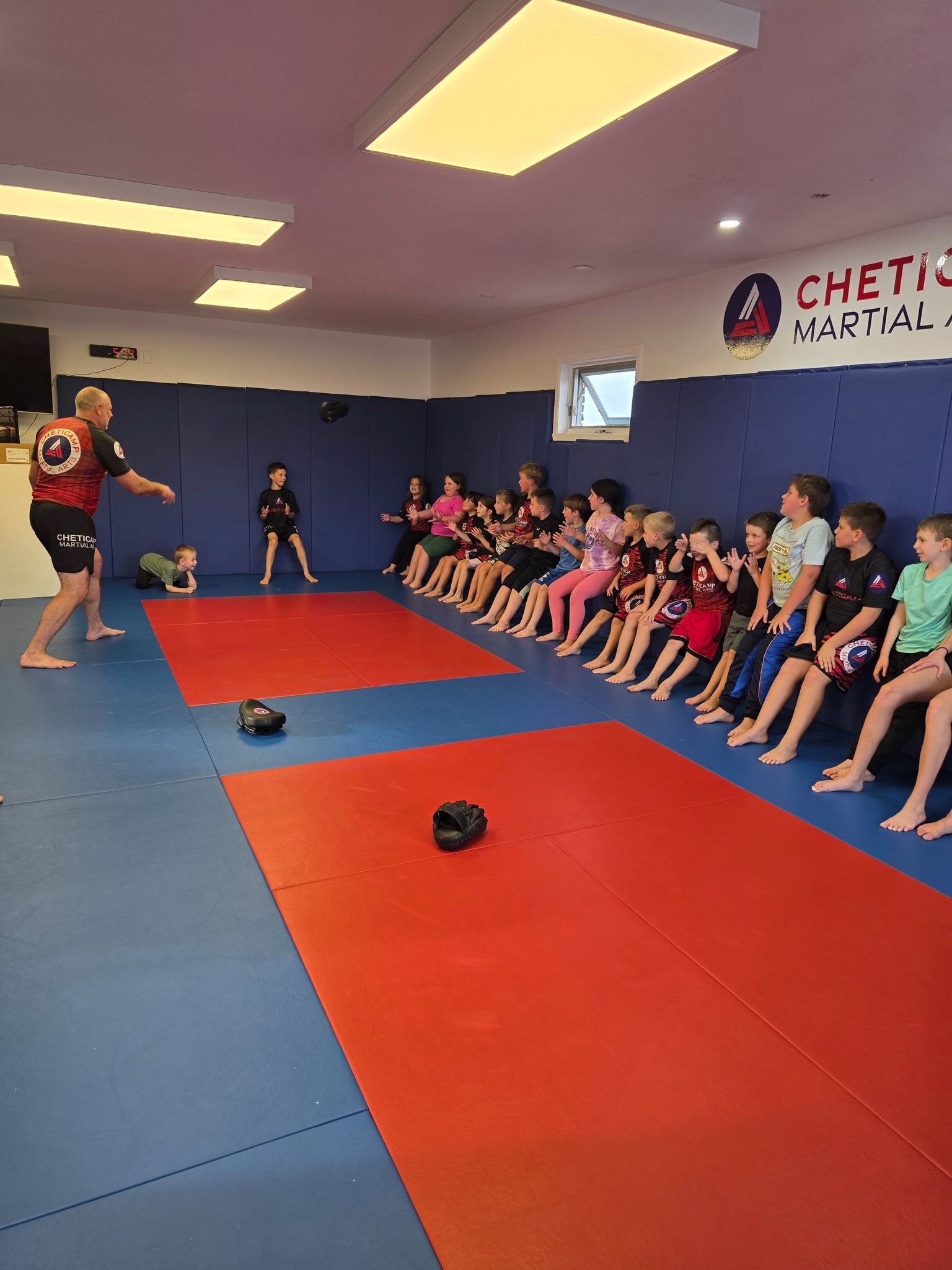Martial arts class on red mats, instructor demonstrating while students sit along the wall.